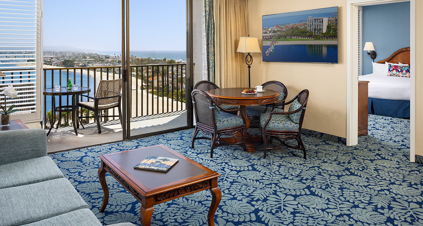 A wide view of a hotel room with a balcony, dining area, sofa, and bed.