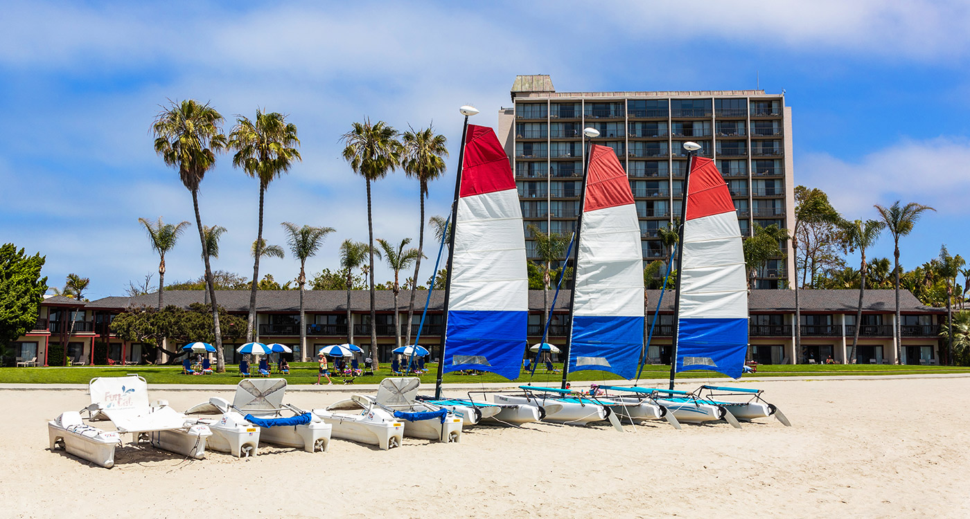 A beach with sailboats and palm trees in front of a hotel building.