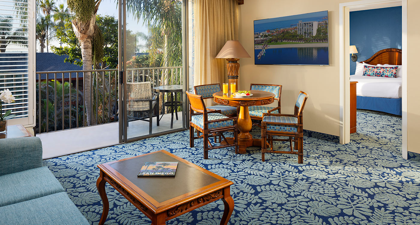A wide view of a hotel suite with a balcony, living area, dining table, and bedroom.