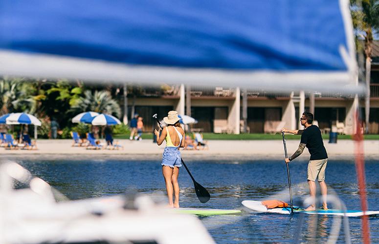 Two people paddleboarding in the water near a beach with lounge chairs and umbrellas.