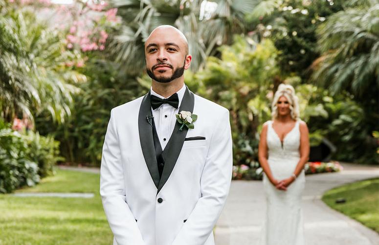 The groom looking sharp in his white tuxedo, with the bride in the background who is stunning in her wedding dress.