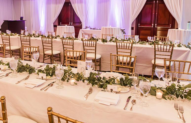 Two rectangular table settings featuring white linens with classic gold framed chairs and greenery on the tables.
