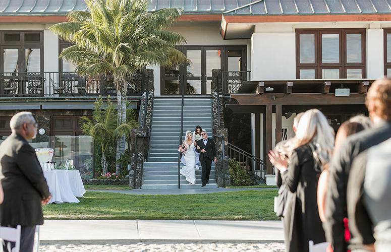 A wedding party is shown walking up the stairs in front of a building with a palm tree.