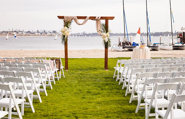 A beachside wedding setup with a wooden arch, white chairs, and sailboats in the background.